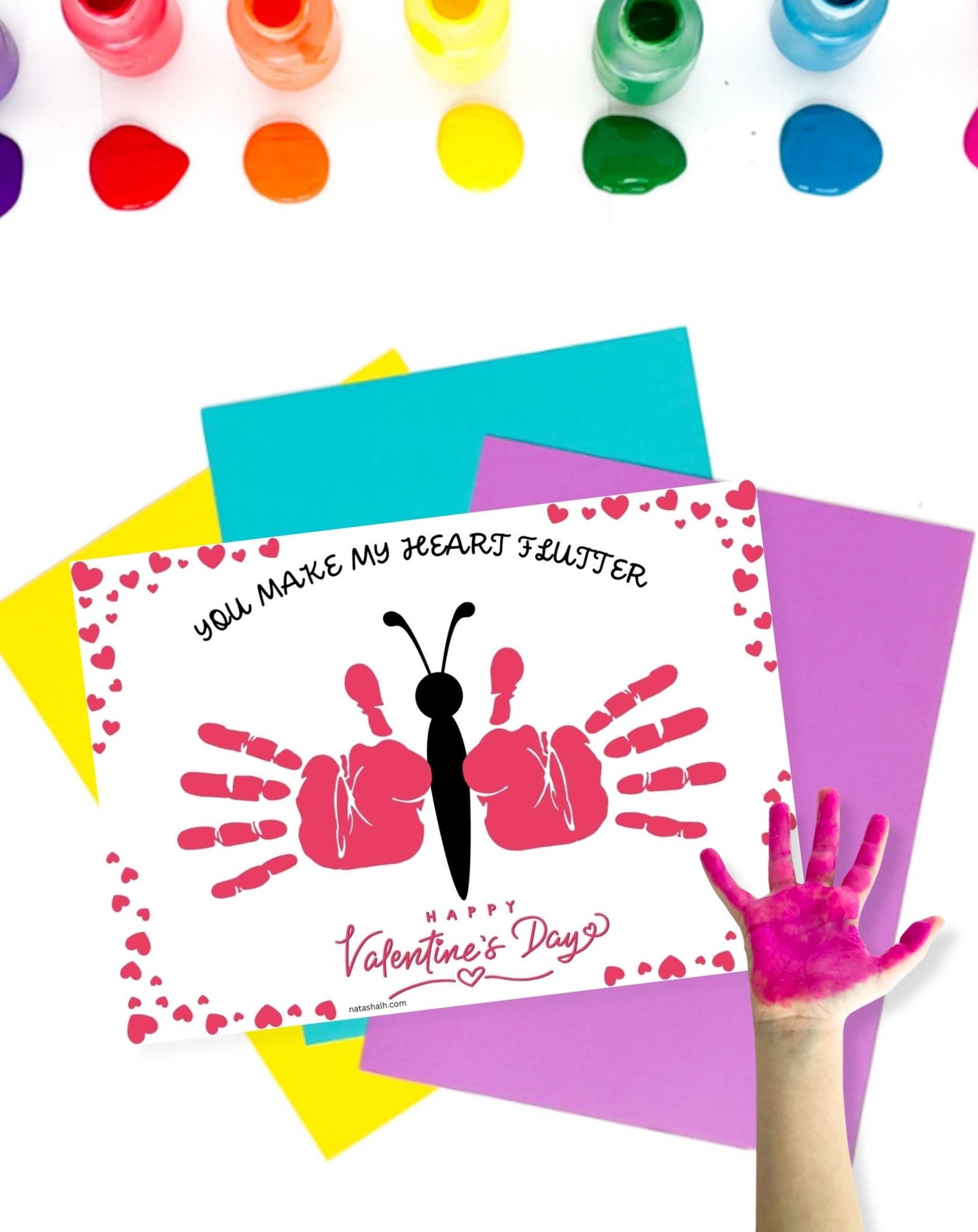 A child's hand with pink paint rests beside a colorful Valentine's Day card featuring handprint butterflies and the text "You Make My Heart Flutter," set against a background of craft supplies for Valentine’s Day handprint crafts.