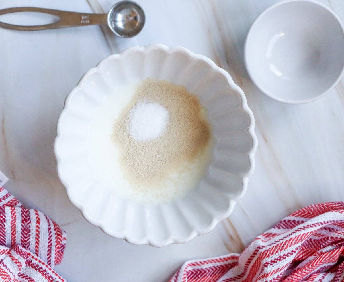 A white bowl with milk sits on a marble surface next to a small bowl of yeast and a red striped kitchen towel, providing ingredients for easy small batch cinnamon rolls or a Valentine's Day cinnamon roll recipe.