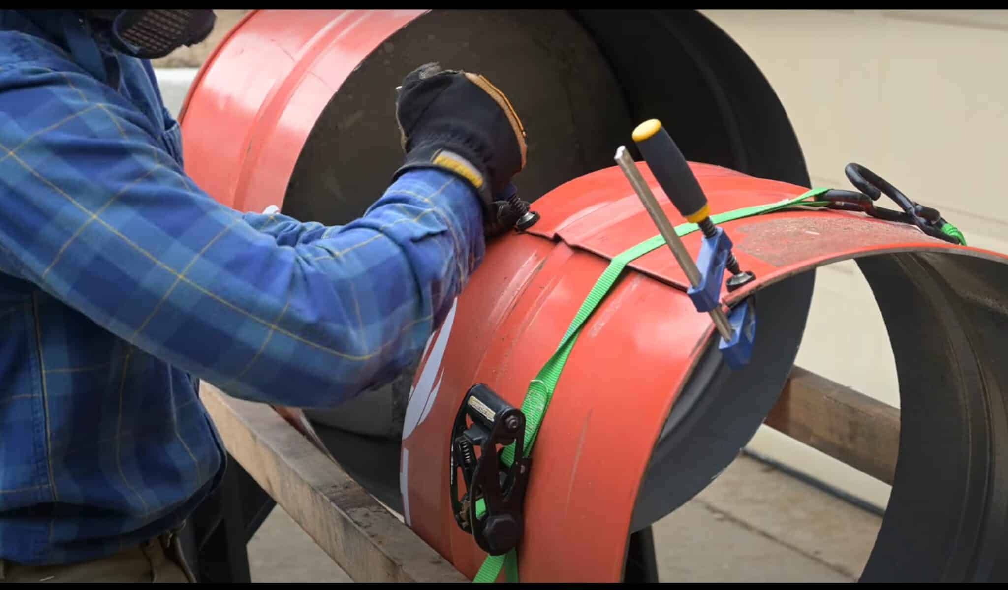 Photo is a of a man folding a 55 gallon drum to reduce the diameter of the barrel.