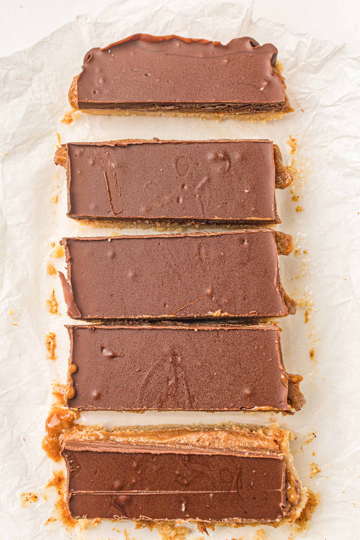 A series of five homemade Twix bars with a chocolate coating laid out on parchment paper, featuring layers of caramel and a base made with almond flour.