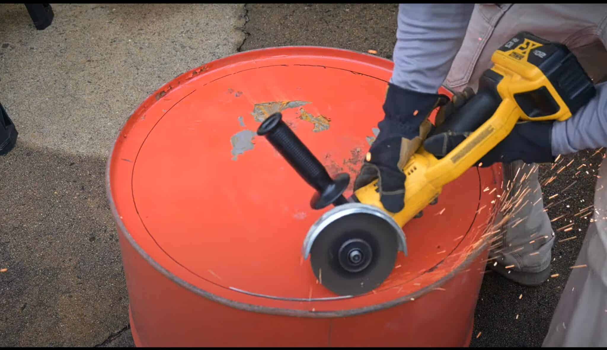 Photo is a man cutting a circular pattern using an angle grinder on the end of a 55 gallon drum.