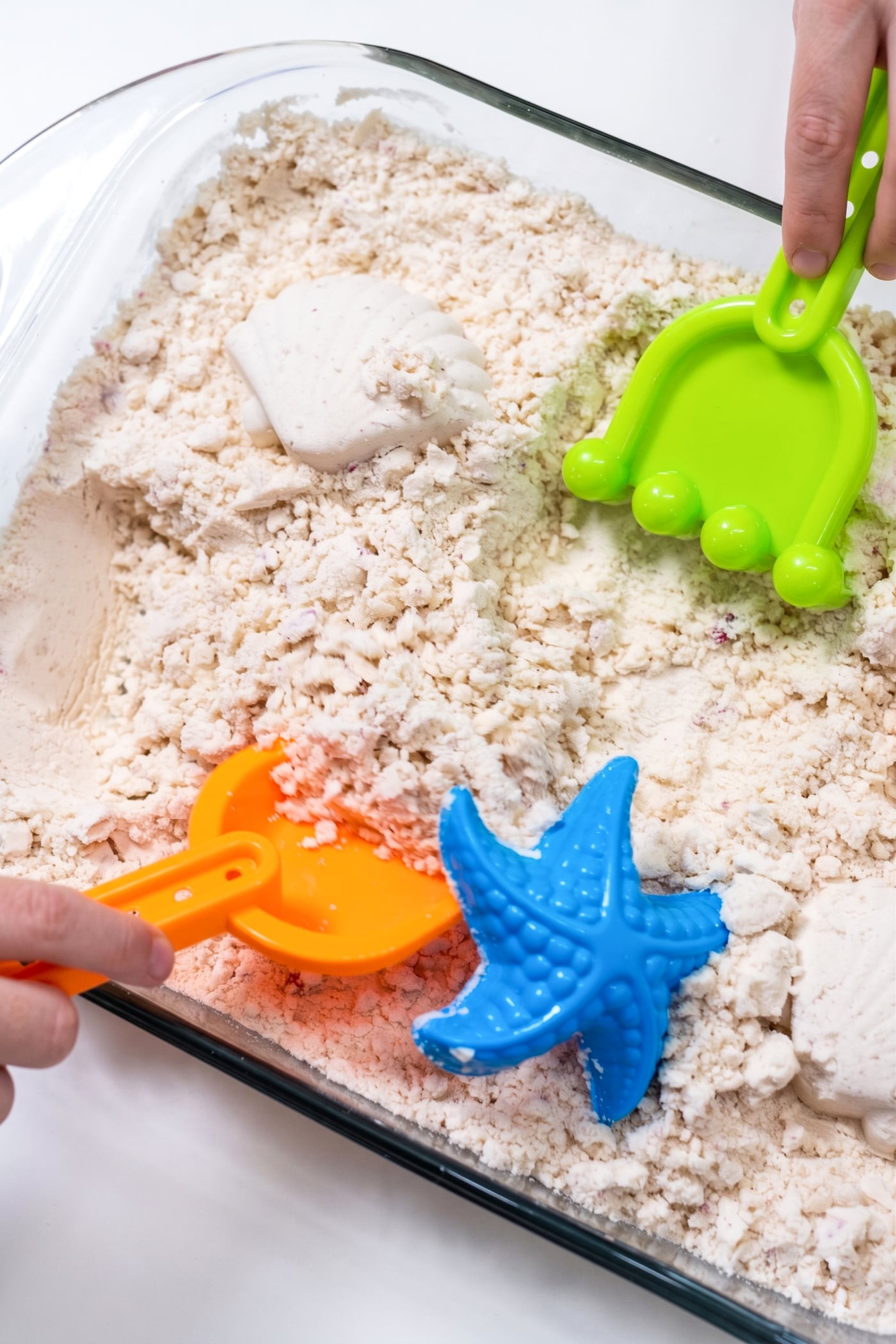 a child's hand with a small plastic rake playing in a dish of kinetic sand
