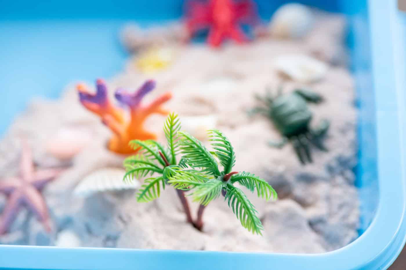 photo of a blue plastic bin with homemade moon sand and plastic items like palm trees