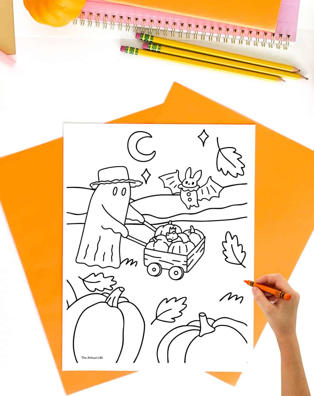 a hand with an orange crayon preparing to color a coloring sheet of a ghost in a pumpkin patch