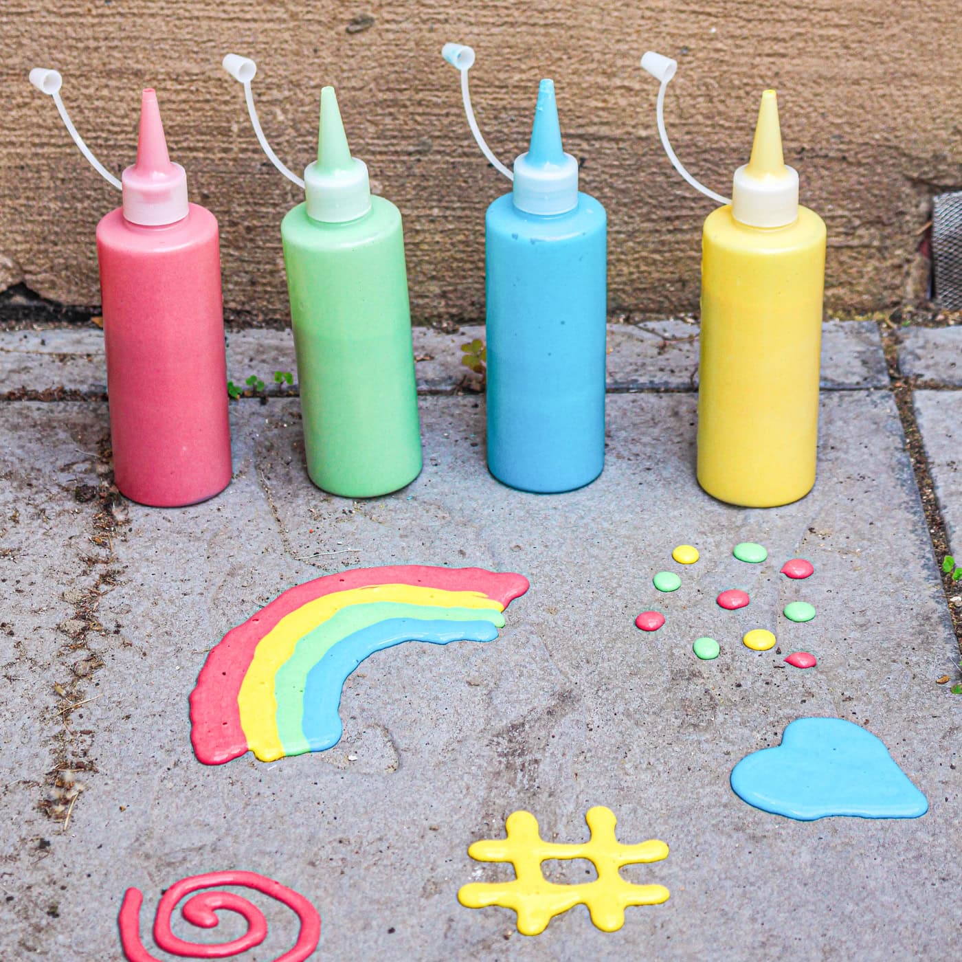 four bottles of sidewalk puff paint in squeeze bottles on the sidewalk in front of a rainbow, dots, a heart, a spiral, and a hash sign made with the puff paint
