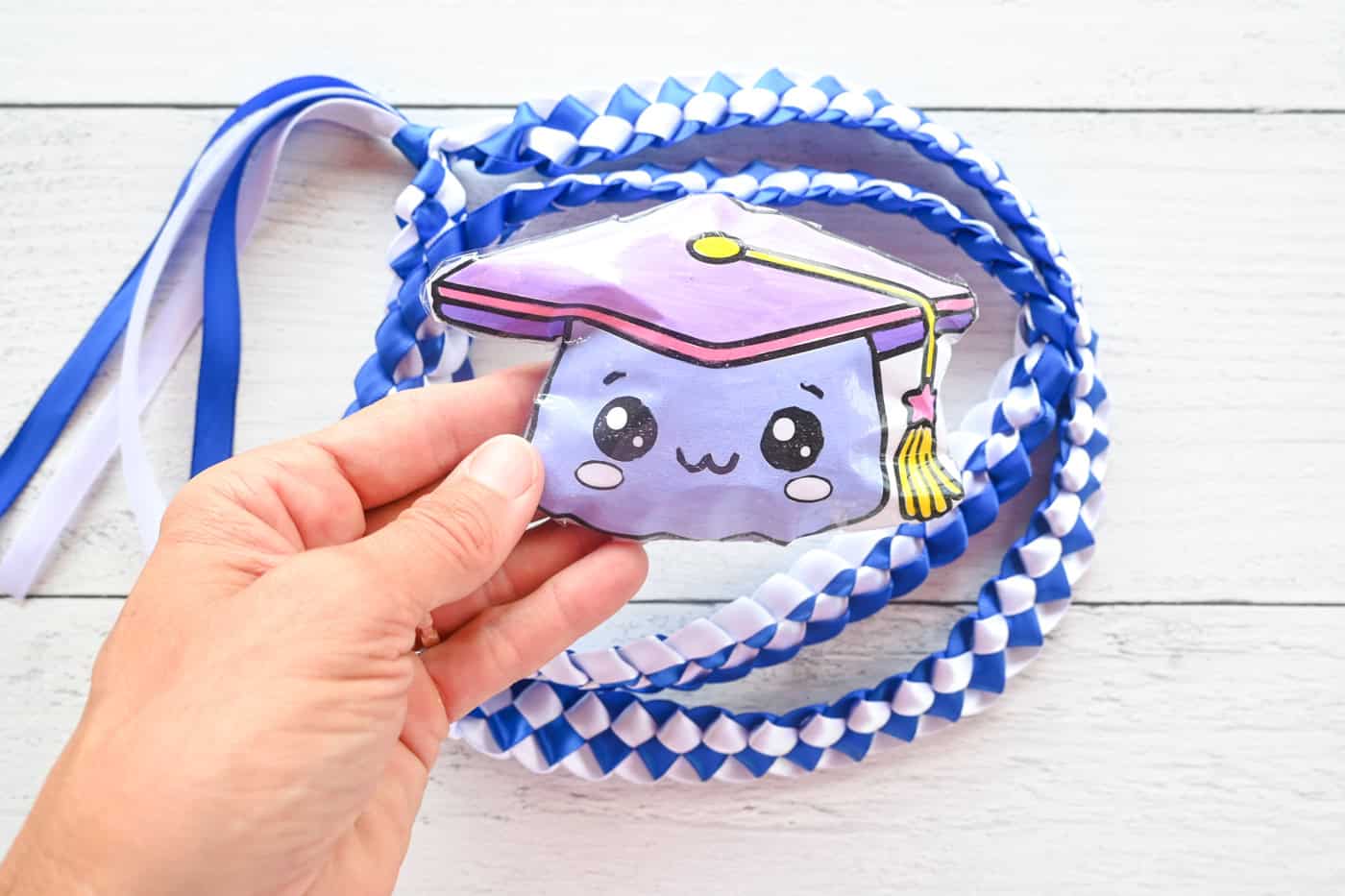 a hand holding a diy graduation cap squishy with a blue and white ribbon lei in the background