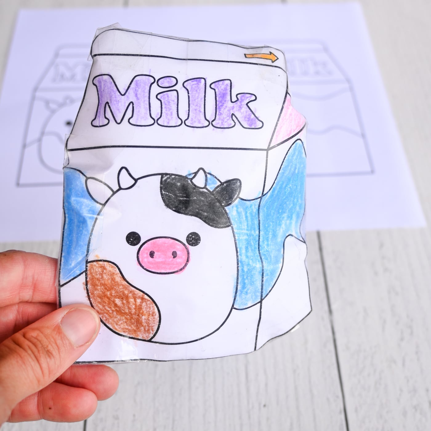 a hand holding a cute milk carton paper squishy in front of a printable paper template for making a IDY paper squishy toy