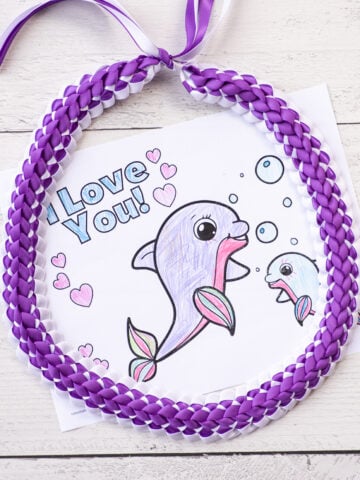 a top down photo of a purple and white double braided Hawaiian ribbon lei with a completed coloring page for Mother's Day