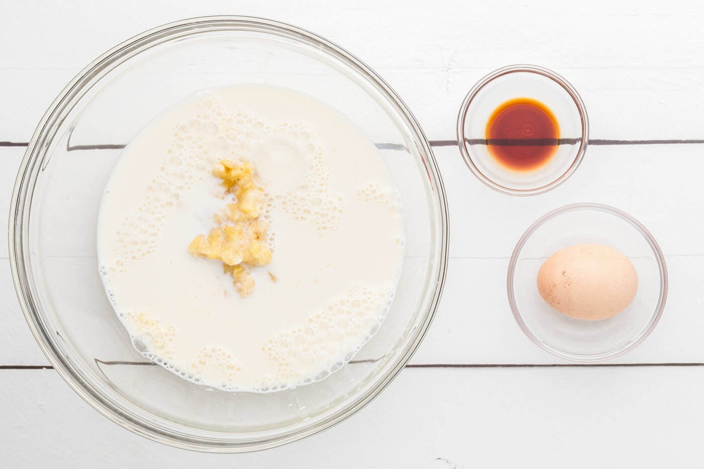 milk, bananas, and vanilla mixed I a bowl next to an egg
