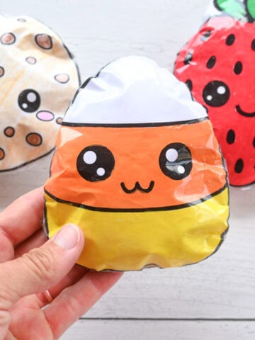 a hand holding a candy corn DIY paper squishy. A cookie and strawberry squishy are visible in the background