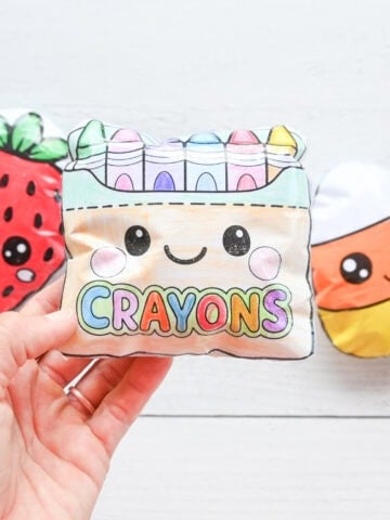 a hand holding a box of crayons paper squishy with a paper strawberry and a paper candy corn in the background