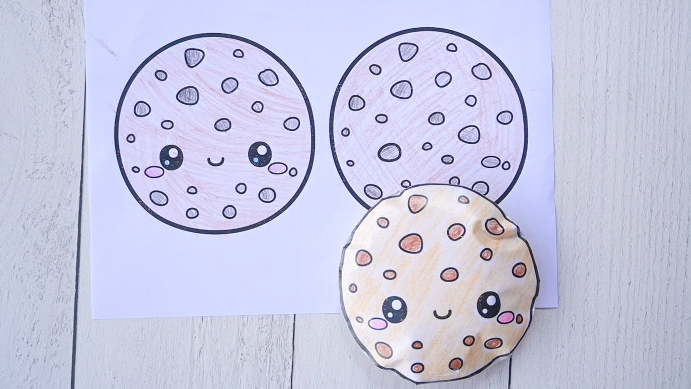 a completed paper squishy of a chocolate chip cookie in front of a colored template page for a chocolate chip cookie squishy
