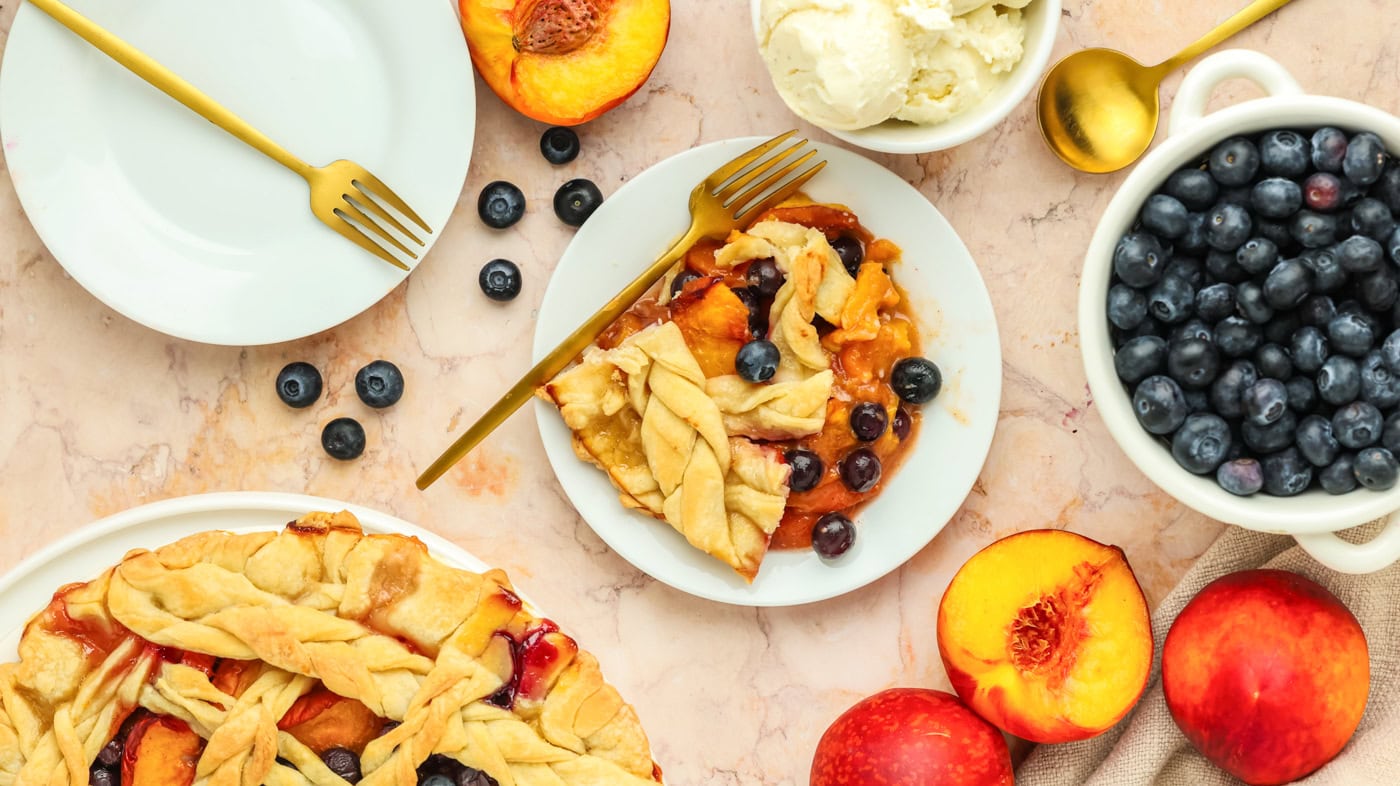 a top down photo of a piece of peach blueberry on a peach marble countertop with fresh peaches and blueberries