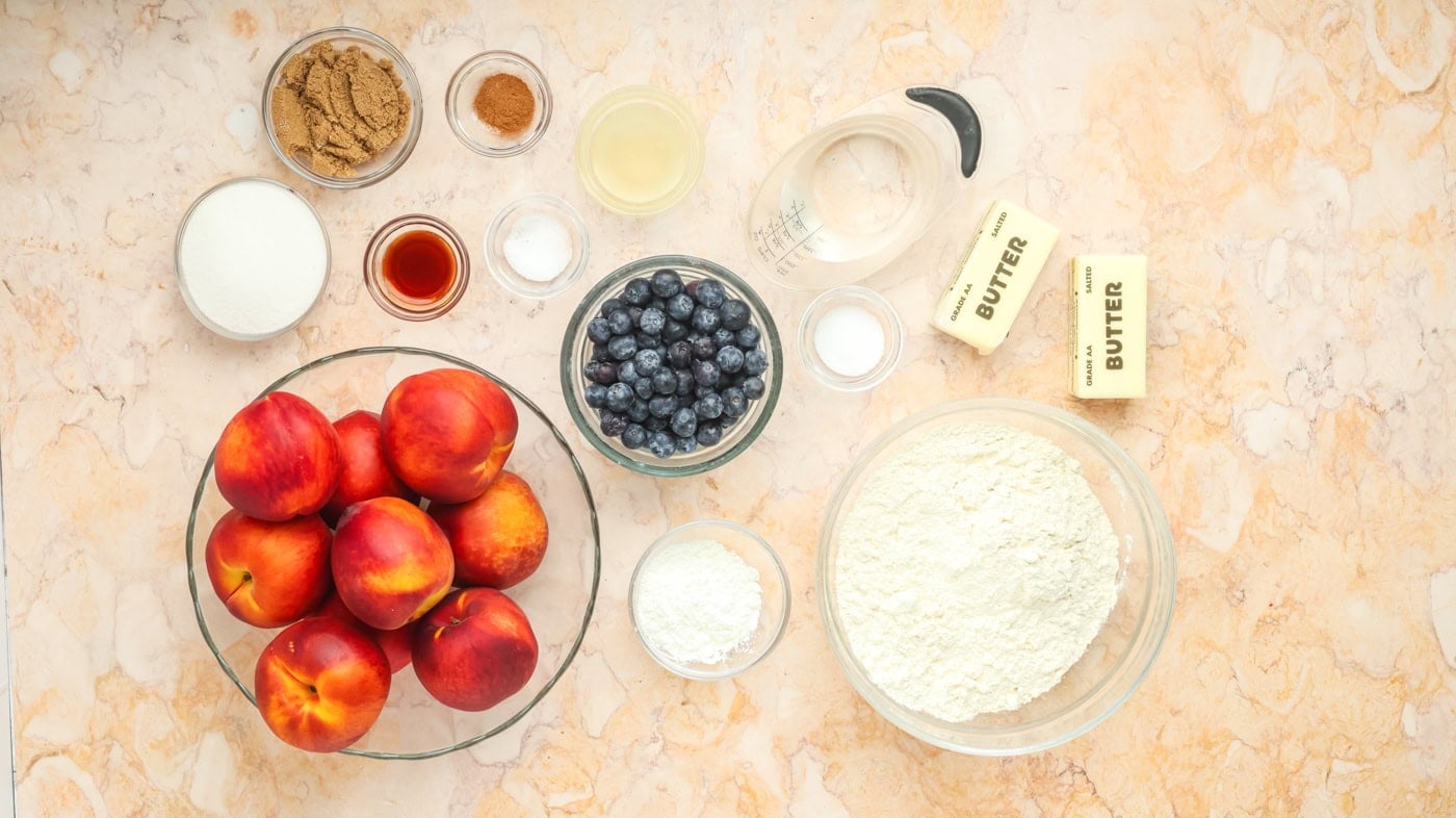 a top down photo of ingredients for a peach pie and crust, including fresh peaches, butter, blueberries, flour, and water