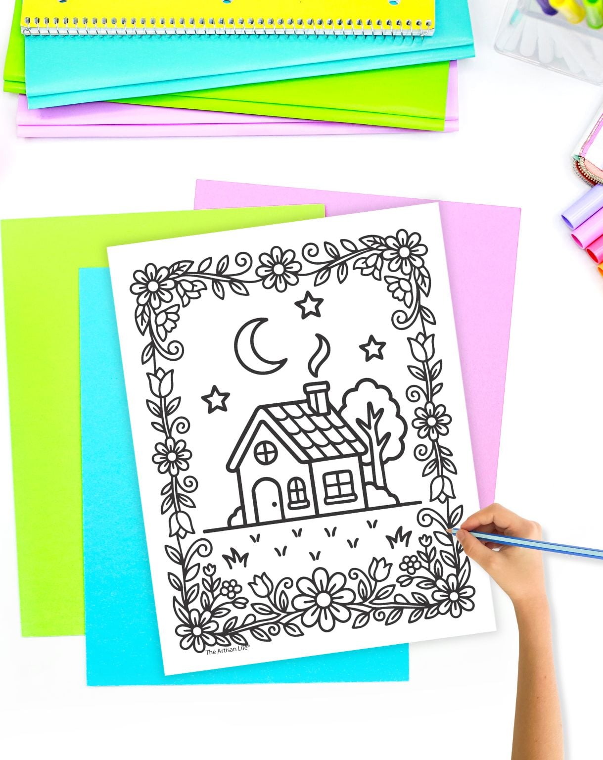 a hand with a blue colored pencil coloring a sheet showing a cute cottage with a floral border