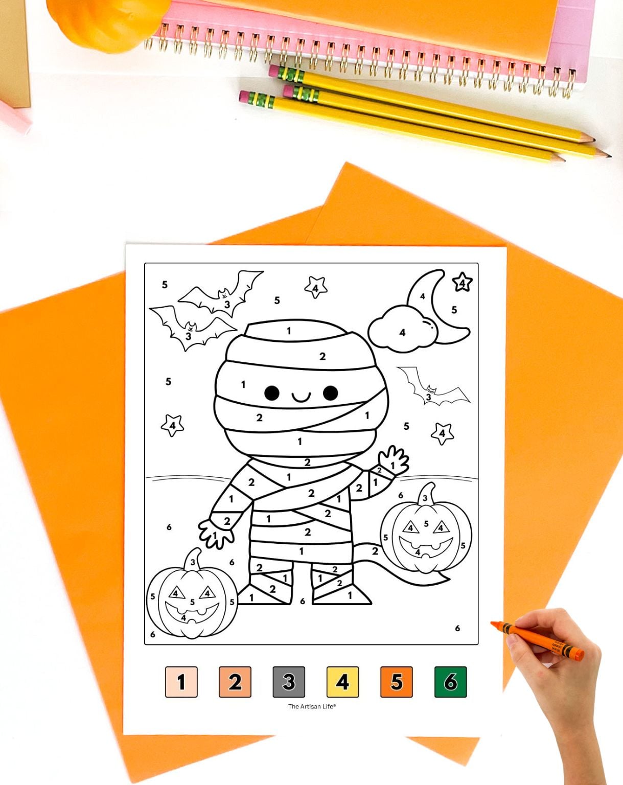 a hadn't with an orange crayon preparing to color a halloween color by number page with a cute mummy