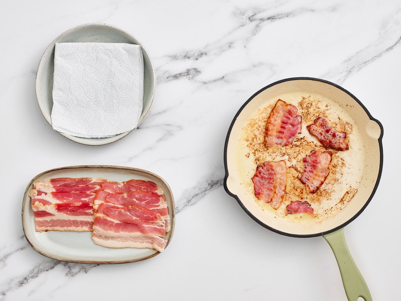 a top down photo of a small skillet cooking bacon next to a plate with paper towels