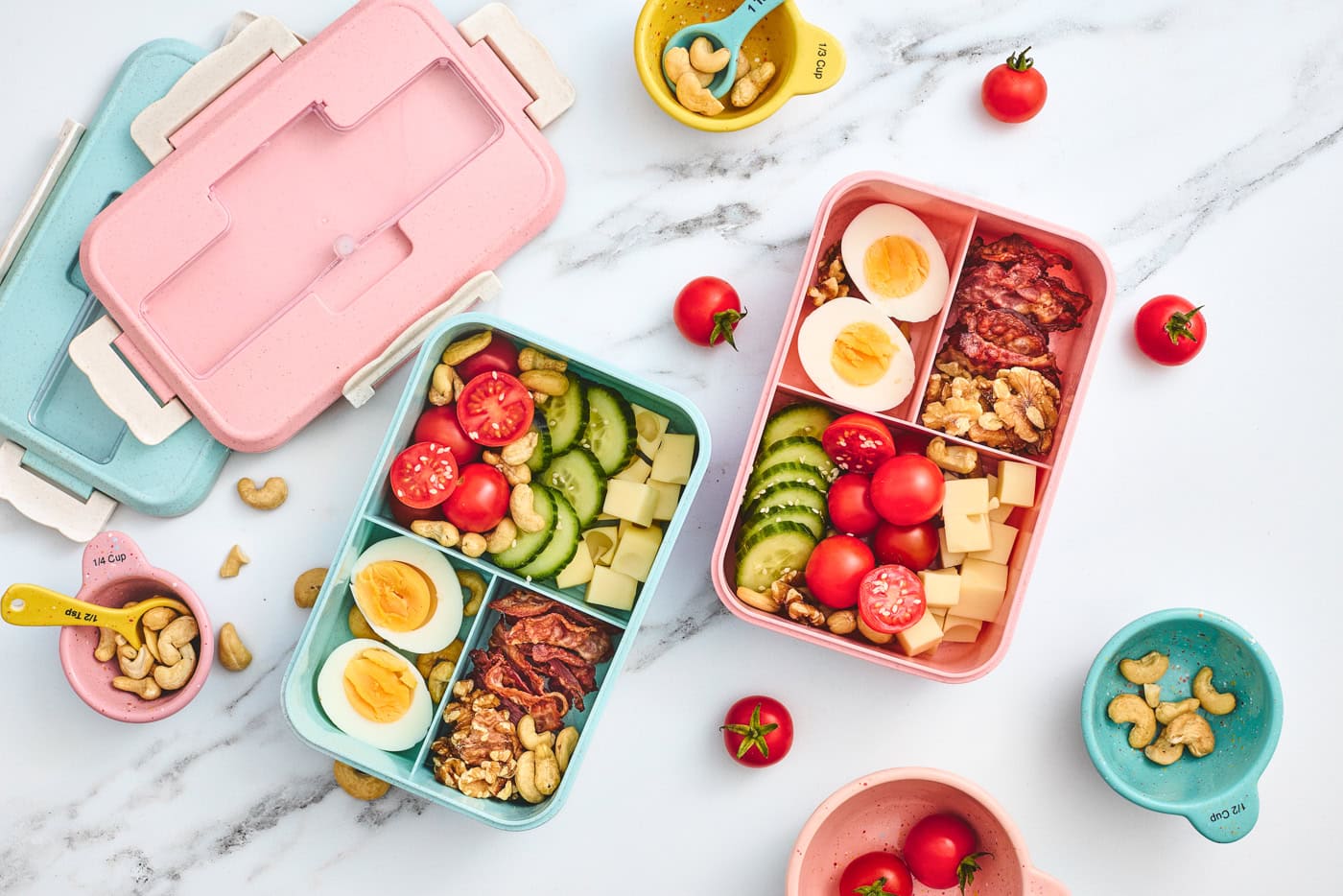 two filled bento style lunchboxes with eggs, veggies, cheese, nuts,a nd bacon