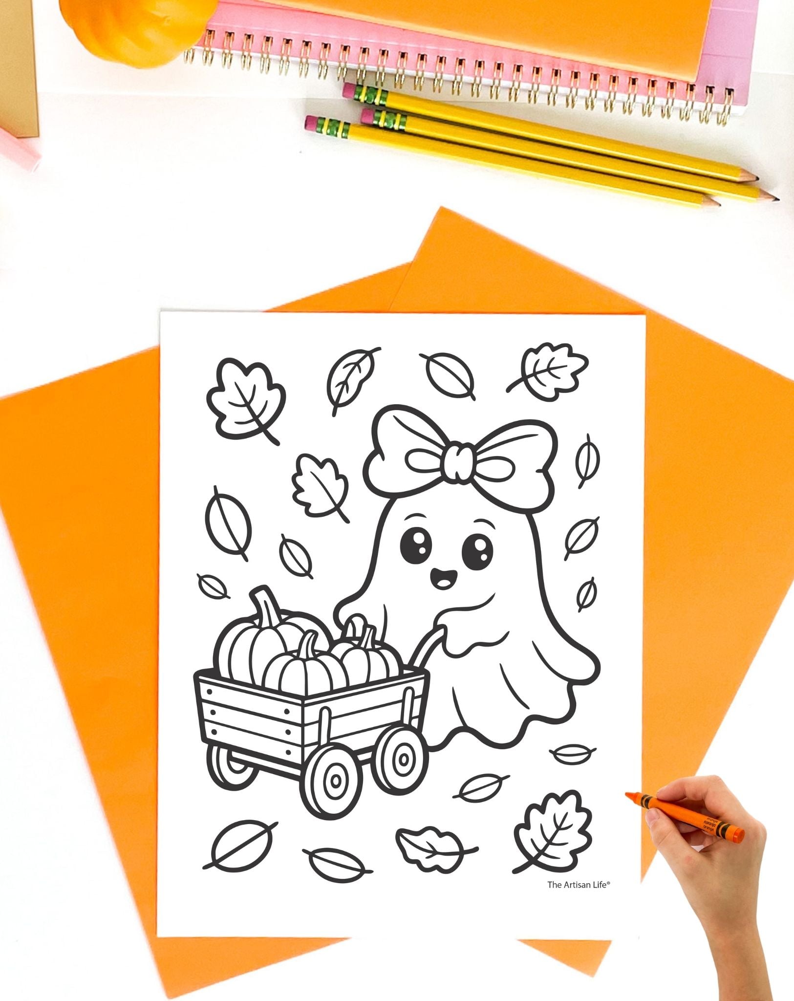 a child's hand with an orange crayon preparing to color a cute ghost coloring page showing a ghost with a wagon of pumpkins