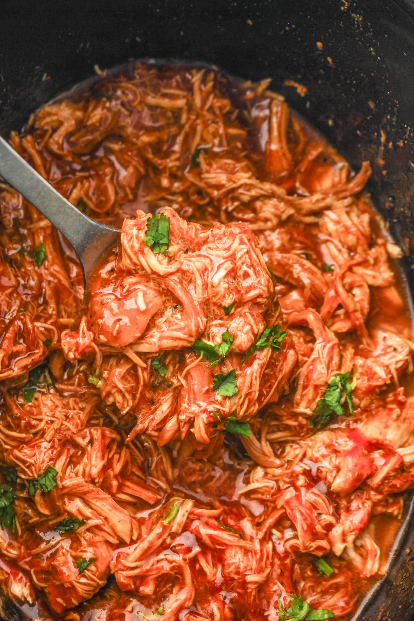 a top down photo of shredded slow cooker honey bbq chicken in a slow cooker crock. A spoon is lifting some of the meat