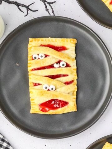 a mummy puff pasty for halloween with eyeballs to look like a mummy. It is on a black plate.