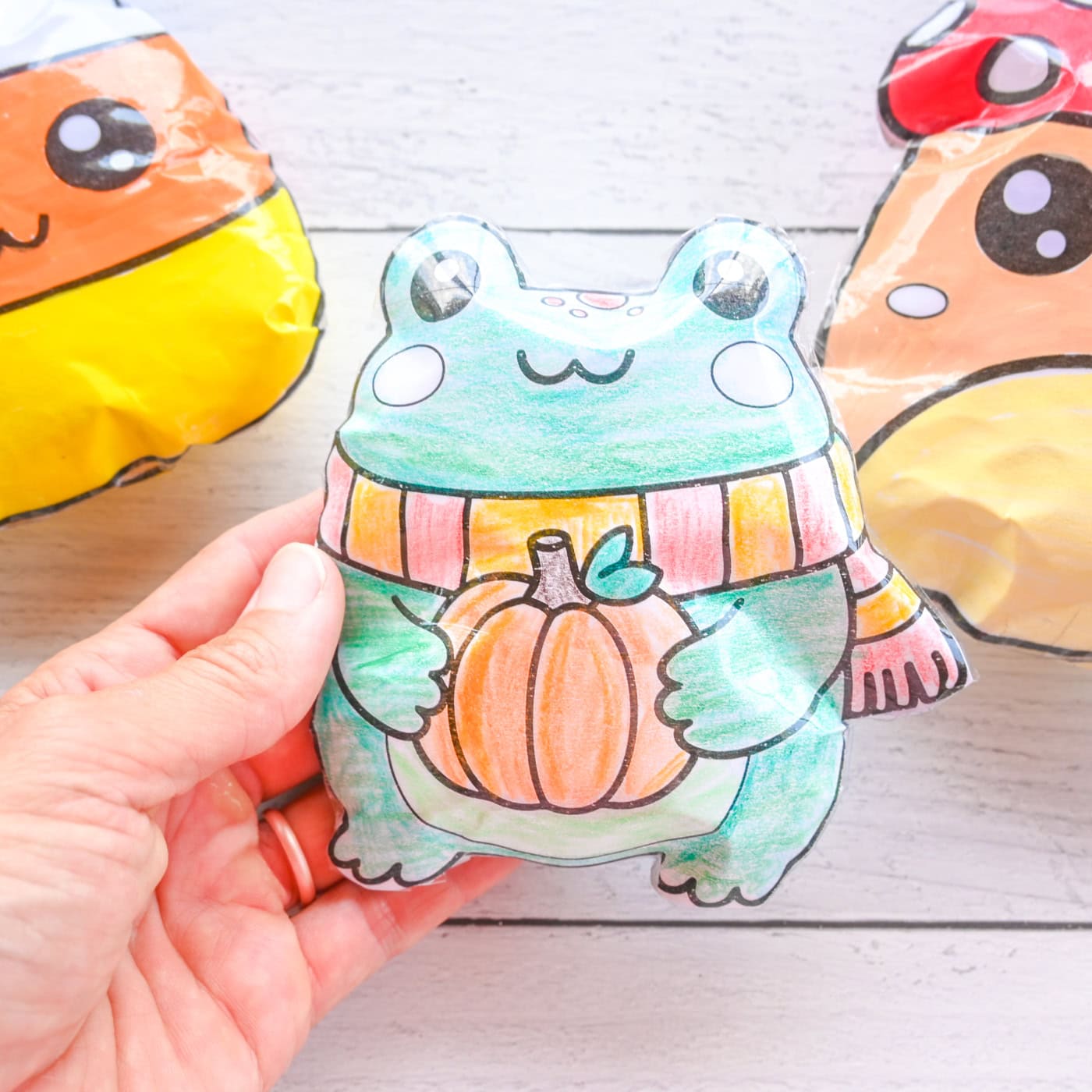 a hand holding a paper frog squishy. A mushroom and candy corn squishy are seen in the background