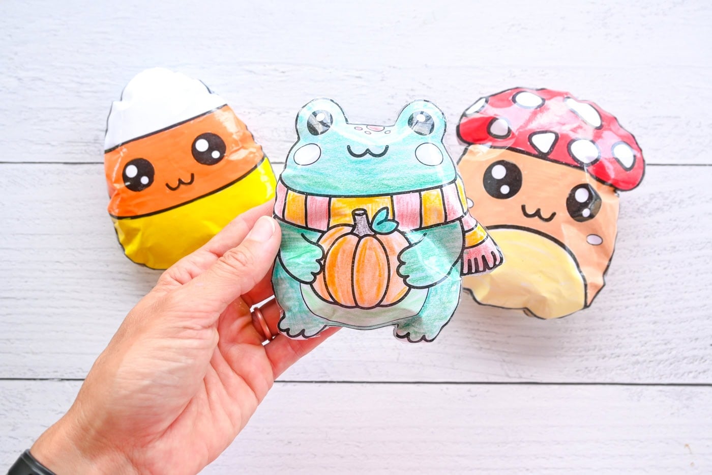 a hand holding a paper frog squishy. A mushroom and candy corn squishy are seen in the background