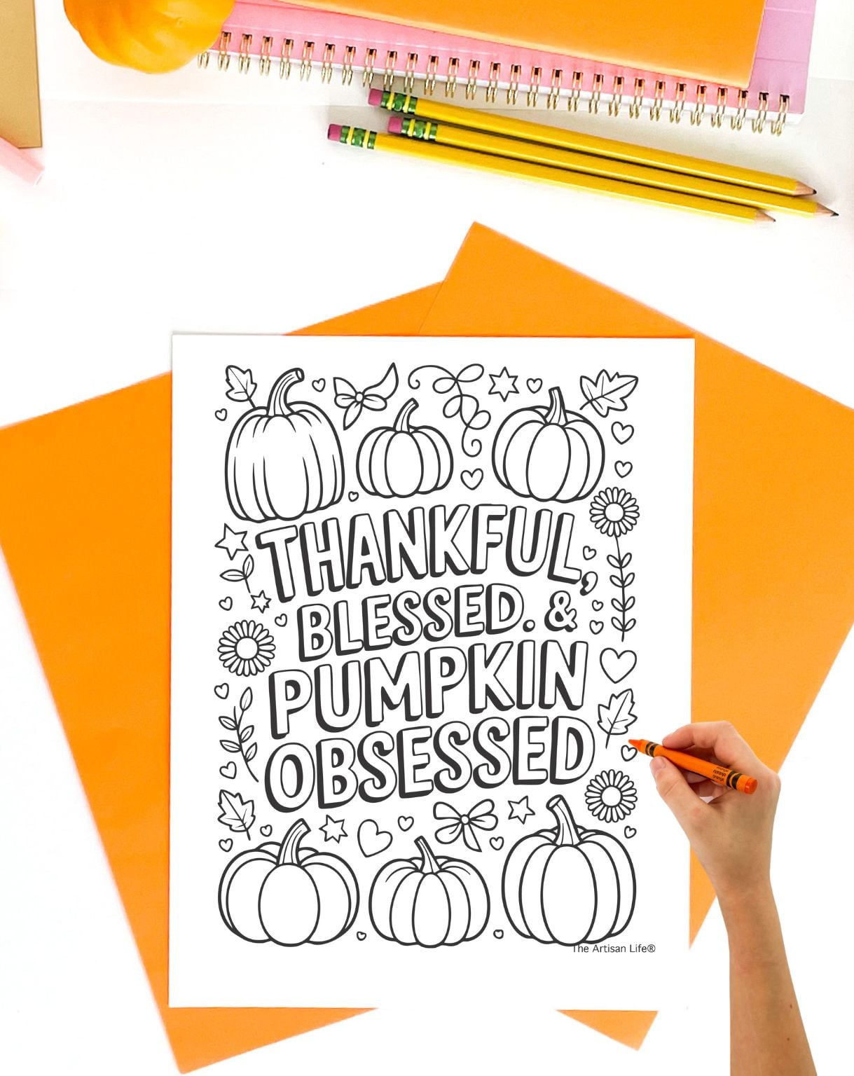 a hand with an orange crayon preparing to color a coloring page with pumpkins, sunflowers, and the text "thankful, blessed, and pumpkin obsessed"